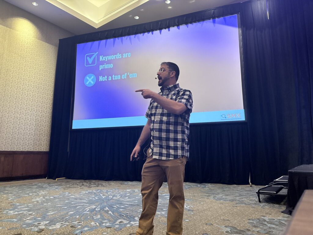 Mordy Oberstein in a plaid shirt stands and gestures while presenting in front of a screen displaying text about keywords at a conference.